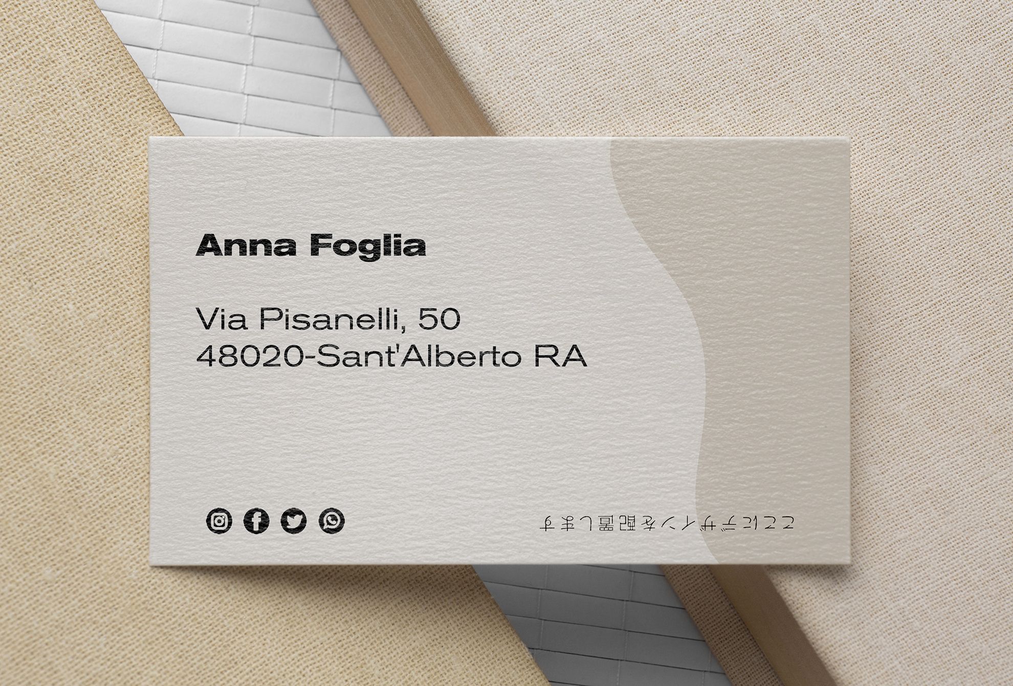 Un biglietto da visita bianco su carta martellata per Anna Foglia con design a onda, indirizzo e icone social media — su superficie tessile beige. I biglietti da visita accattivanti su carte speciali con texture martellata o vergata aggiungono qualità tattile premium a qualsiasi brand personale o identità professionale.