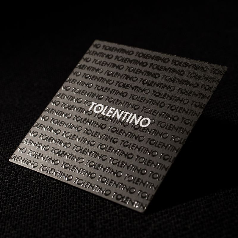 Un biglietto da visita quadrato nero con vernice UV tono su tono in rilievo con motivo ripetuto del nome 'Tolentino' e logo centrale in bianco. I biglietti da visita eleganti quadrati con vernice UV o goffratura tono su tono sono ideali per brand di moda, luxury e designer che desiderano una carta tattile ad alto impatto.