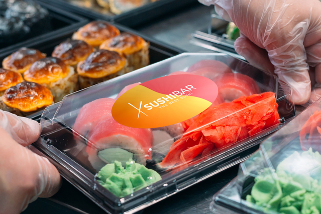 Un adesivo ovale in vinile arancione brillante con logo del brand sushi applicato su un contenitore trasparente per cibo da asporto. L'adesivo spicca vivacemente sui sushi colorati e le mani con guanti che preparano l'ordine. Gli adesivi ovali in vinile personalizzati Sprint24 sono perfetti per il packaging alimentare, il branding e l'etichettatura promozionale.