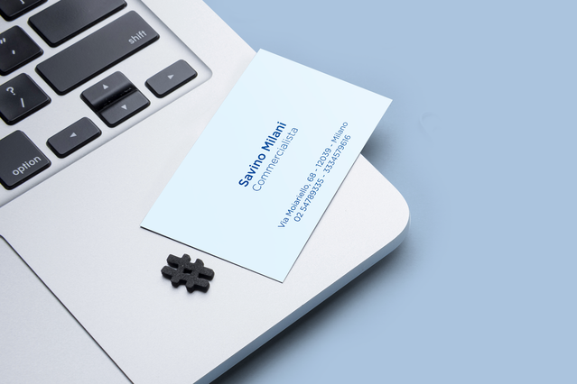 Un biglietto da visita verticale azzurro per 'Savino Millani, Commercialista' appoggiato contro la tastiera di un laptop su sfondo blu tenue, con un piccolo dettaglio puzzle hashtag. I biglietti da visita personalizzati per commercialisti in tonalità pastello moderne proiettano un'identità professionale fresca e accattivante — ideali per consulenti fiscali e finanziari che vogliono rinnovare il p…