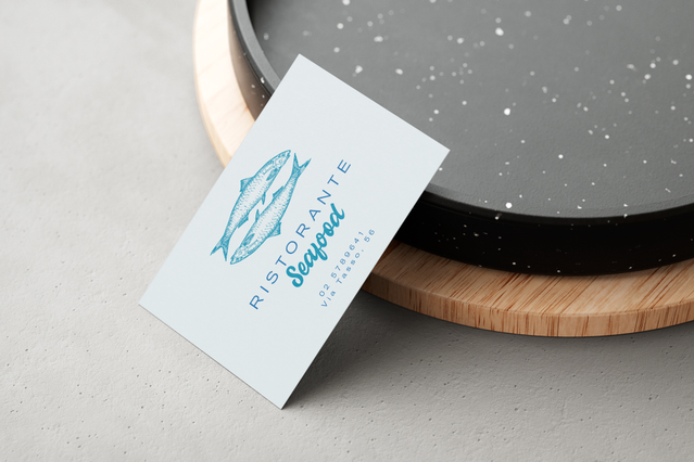 Un biglietto da visita ristorante verticale bianco per 'Ristorante Seafood' con logo illustrazione pesce teal, fotografato su un vassoio di servizio rotondo in legno con sfondo in pietra scura. I biglietti da visita personalizzati per ristoranti uniscono un design elegante alle informazioni di contatto — ideali da lasciare sui tavoli, alla reception o da includere negli ordini d'asporto per fidel…