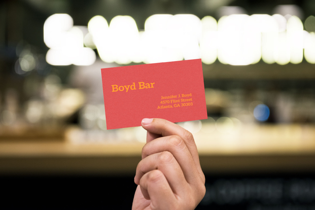 Un biglietto da visita 'Boyd Bar' rosso brillante stampato su carta colorata Sirio Fedrigoni con testo giallo, tenuto davanti allo sfondo di un bar illuminato. Sprint24 stampa biglietti da visita in carta colorata in 6 tonalità Sirio vivaci con opzione stampa a caldo — perfetti per bar, ristoranti e brand creativi.