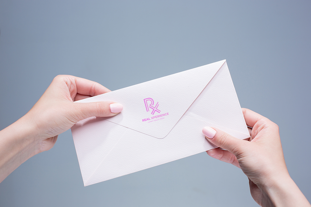 Le mani di una donna che tengono una busta bianca stampata con un logo rosa "RX" su sfondo grigio morbido. Questa immagine illustra la stampa personalizzata di buste con logo di Sprint24, ideale per aziende, studi medici e professionisti che desiderano materiali postali brandizzati. Le buste personalizzate aggiungono un tocco premium alla tua corrispondenza.