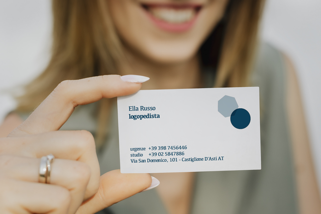 Una donna sorridente tiene in mano un biglietto da visita personalizzato per logopedista con il nome Ella Russo e dettagli di contatto su un design bianco pulito con accenti a cerchi teal. Il biglietto dimostra la qualità di stampa professionale ideale per logopedisti che vogliono lasciare un'impressione duratura. Stampati online con Sprint24, questi biglietti da visita per logopedista sono prati…