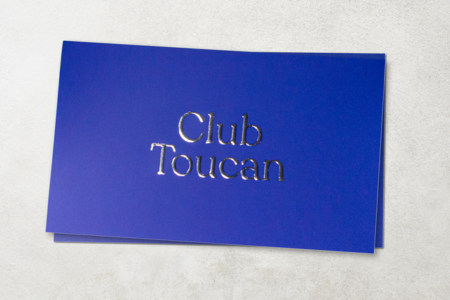 Due biglietti da visita blu cobalto con 'Club Toucan' in tipografia serif goffrata argento, impilati su superficie bianca testurizzata. I biglietti da visita premium con stampa a caldo oro e argento e lettering goffrato creano un'impressione tattile lussuosa — ideali per club esclusivi, locali di alto livello e brand che cercano finiture metalliche sofisticate.