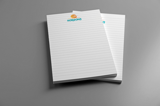 Due block notes A4 personalizzati con template a righe e logo colorato del brand di viaggio Horizons in cima su superficie grigia. I block notes A4 personalizzati con template a righe, quadretti, puntinato o millimetrato e branding aziendale sono articoli di cancelleria promozionale ideali per agenzie di viaggio, aziende e professionisti da distribuire a riunioni, eventi e come gadget corporate.