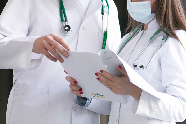 Due medici in camice bianco con stetoscopi verdi che consultano una cartellina porta referti brandizzata in un corridoio clinico. Sprint24 stampa cartelline porta referti medici personalizzate con fustella e logo per medici, ospedali e studi privati — ideali per consegnare referti in modo professionale.
