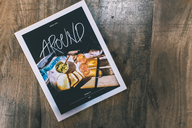 Un flyer pubblicitario stampato per la rivista 'Around' con copertina di fotografia food e tipografia scritta a mano in grassetto su superficie in legno. I flyer pubblicitari personalizzati sono uno strumento di marketing stampato ad alto impatto ideale per brand food, ristoranti, riviste, eventi e lanci di prodotto — stampabili fronte/retro per massimo impatto del messaggio a basso costo.