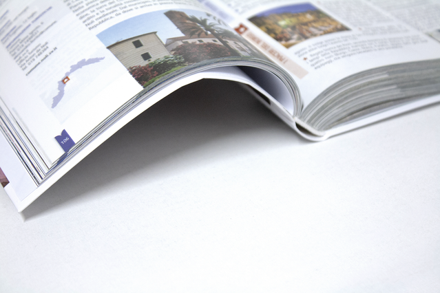 Un libro cartonato flessibile con rilegatura olandese aperto che mostra una doppia pagina multicolonna con fotografie di viaggio, foto di edifici e monumenti e colonne di testo editoriale, su sfondo bianco. I libri con rilegatura olandese (brossura olandese) combinano la solidità di un cartonato con la flessibilità e la leggerezza di una brossura — ideali per guide turistiche, manuali, libri azie…