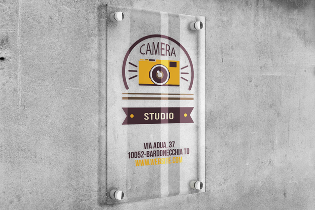 Un'insegna in plexiglass trasparente personalizzata 'Camera Studio' con logo fotocamera vintage e indirizzo, montata su muro di cemento con distanziali cromati. Ideale per studi fotografici, negozi e uffici professionali che cercano insegne in plexiglass eleganti e brandizzate (30x40cm fino a 110x210cm, 3 o 5mm) che resistono alle intemperie per oltre 10 anni e aumentano la visibilità su strada.