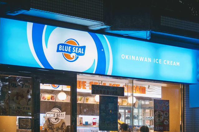 Una vivace insegna luminosa in plexiglass opalino retroilluminata blu 'Blue Seal Okinawan Ice Cream' montata sopra l'ingresso di un negozio, che brilla brillantemente nella notte. Ideale per negozi, ristoranti e attività commerciali che cercano insegne luminose su plexiglass opalino personalizzate (3 o 5mm, stampa fronte-retro) per massima visibilità su strada e rafforzamento dell'identità del br…