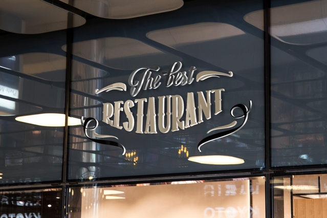 Una vetrina di ristorante mostra eleganti lettere tridimensionali dorate che scrivono 'This Is RESTAURANT' sul vetro. Le lettere in rilievo creano profondità con ombre visibili sul vetro. Questi cartelli 3D professionali in forex, plexiglas o alluminio Dibond® sono perfetti per ristoranti, negozi e uffici.
