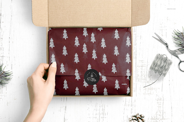 Una mano che apre una scatola di cartone kraft per rivelare carta da imballo bordeaux personalizzata con motivo alberi di Natale argento e sigillo di ceralacca brandato, circondata da rametti di pino e nastro. Ideale per boutique, gioiellerie, pasticcerie e artigiani che cercano carta da imballo personalizzata premium per trasformare ogni presentazione prodotto in un'esperienza regalo elegante e …