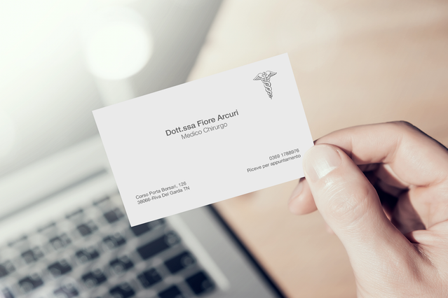 Una mano che tiene un biglietto da visita bianco per 'Dott.ssa Flora Accuri, Medico Chirurgo' con logo caduceo medico, vicino a una tastiera di laptop. I biglietti da visita personalizzati per medici e chirurghi rafforzano la credibilità professionale e la fiducia dei pazienti — completamente personalizzabili con nome, specialità, contatti e logo medico.