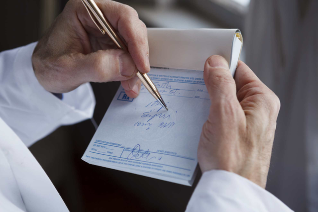 Un medico in camice bianco che scrive su un blocco ricettario medico personalizzato tenuto in mano, con penna dorata. Sprint24 stampa blocchi ricette mediche personalizzati con intestazione per medici, MMG, veterinari e cliniche — ideali per ricettari professionali brandizzati con recapiti e logo dello studio.