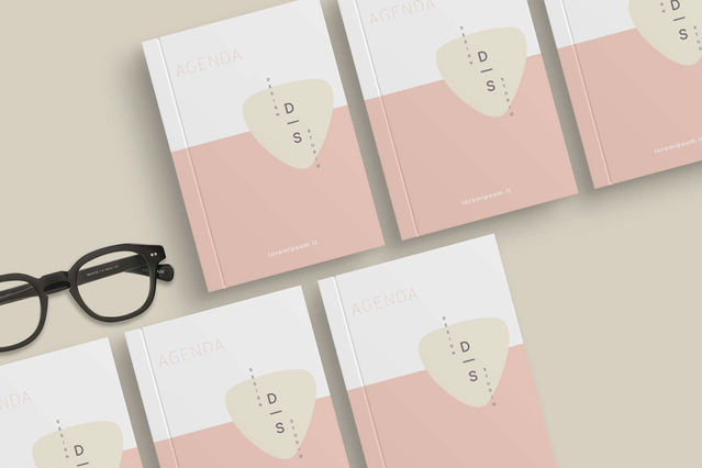 Cinque agende personalizzate con rilegatura in filo refe con elegante copertina minimal rosa e crema con branding 'D|S' e URL sito, disposte su superficie beige accanto a occhiali neri. Sprint24 stampa agende in filo refe personalizzate in vari formati — ideali per aziende e professionisti che cercano agende brandizzate durature e di alta qualità.