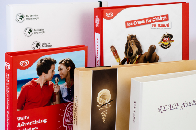 Una pila di raccoglitori ad anelli personalizzati con volumi rossi brandizzati 'Wall's Advertising' e 'Ice Cream for Children PR Manual' con eleganti raccoglitori bianchi 'REALE gioielli' e beige. Sprint24 stampa raccoglitori ad anelli personalizzati per comunicazione aziendale, manuali di brand e kit PR — perfetti per presentazioni vendita e identità corporate.
