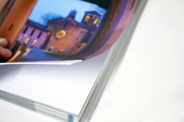Un primo piano di un libro cartonato flessibile con rilegatura olandese aperto delicatamente, che mostra la copertina pieghevole con una foto a tutta pagina di una piazza italiana illuminata la sera con una chiesa. La copertina flessibile dei libri con rilegatura olandese si ripiega completamente, rendendoli leggeri, resistenti e facili da maneggiare — ideali per guide turistiche, libri d'arte, p…