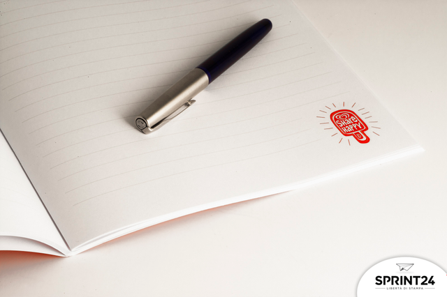 Quaderno a punto metallico premium che mostra pagine bianche brillanti in carta patinata di qualità, ideale per professionisti. La vista interna rivela il branding rosso distintivo, il doppio punto metallico e la plastificazione opaca che caratterizzano questo regalo aziendale e prodotto da ufficio.