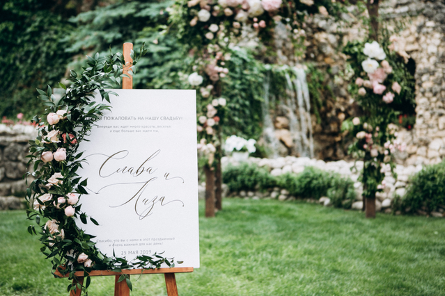 Un tableau matrimonio bianco in calligrafia montato su cavalletto in legno davanti a un'arco floreale rigoglioso in una cerimonia all'aperto. Sprint24 stampa tableau di nozze personalizzati in vari materiali e formati — ideali per organizzare i posti a sedere al ricevimento con nomi e numero tavoli.