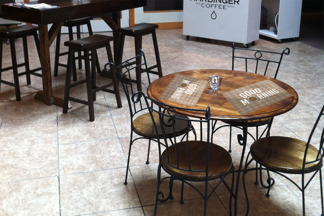 Tovaglietta personalizzata stampata su tavolo in legno in un'ambientazione cafe moderna con sedie nere. Mostra la qualità di stampa professionale idonea per lino da tavola marcato in ristoranti e cafe. Le tovagliette personalizzate migliorano l'atmosfera del servizio e il branding dell'attività ristorazione.