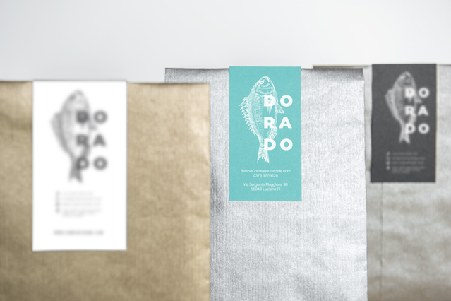 Tre diversi sacchetti di carta — bianco, kraft e grigio — ciascuno sigillato con un'etichetta chiudipacco personalizzata 'Dorado' con logo pesce nei colorway abbinati: bianco, teal e antracite scuro. Ideali per pescherie, attività alimentari e produttori artigianali che cercano etichette chiudipacco adesive coerenti e brandizzate per packaging coordinato.